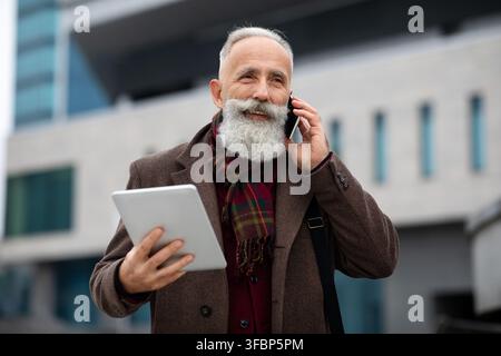 Bell'uomo dai capelli grigi che parla al telefono mentre si va per strada Foto Stock