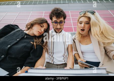 Tre giovani adulti dall'aria sorpresa mentre esaminano documenti all'aperto davanti a un muro colorato Foto Stock