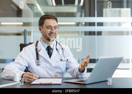 Medico maschio in camice bianco con stetoscopio seduto alla scrivania, sorridendo e spiegando una diagnosi durante una consultazione video di telemedicina su un laptop in un ufficio clinico Foto Stock