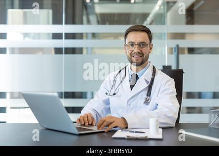Medico maschio sicuro di sé in camice da laboratorio e stetoscopio seduto alla scrivania sorridendo alla telecamera mentre utilizza un laptop in un ufficio medico moderno, per la telemedicina e l'assistenza al paziente Foto Stock