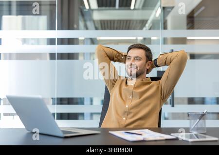 Giovane uomo d'affari che si rilassa con le mani dietro la testa, godendosi un momento di soddisfazione e successo in un ambiente d'ufficio moderno dopo aver completato i suoi compiti Foto Stock