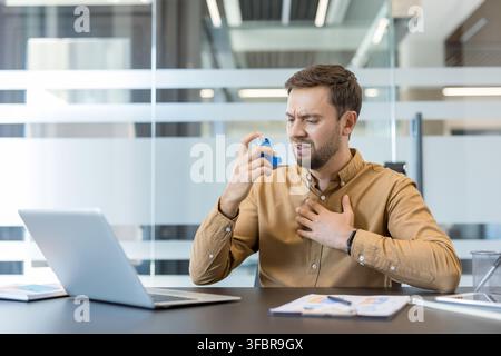 Giovane uomo che usa un inalatore medico blu per l'asma, sperimentando difficoltà respiratorie e dolori al petto mentre è seduto alla sua scrivania in un ufficio moderno, lavorando su un laptop Foto Stock