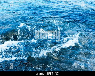 Vista ravvicinata dell'acqua blu profondo dell'oceano con piccole onde, increspature e schiuma bianca che formano una trama naturale. Foto Stock