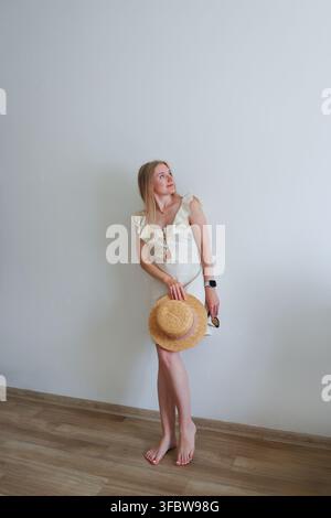 Donna felice che indossa un cappello di paglia e tiene occhiali da sole, concetto di vacanza estiva rilassata e turismo su sfondo minimalista. Foto Stock
