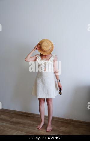 Donna scalza in vestito estivo bianco che copre il viso con un cappello di paglia e tiene occhiali da sole, concetto minimalista di vacanza con spazio per copiare. Foto Stock