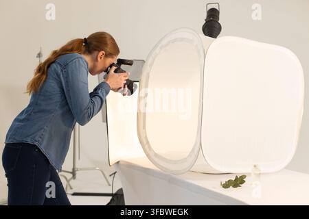 Donna in camicia di jeans appoggiata, usa flash DSLR, scatta bottiglia di profumo in softbox in studio, spazio copia Foto Stock