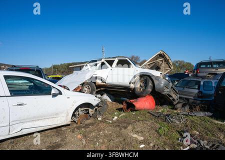 Un'auto bianca danneggiata e un veicolo bianco impilato con il cofano aperto, situati in un centro di smaltimento auto all'aperto contro un cielo blu. Foto Stock