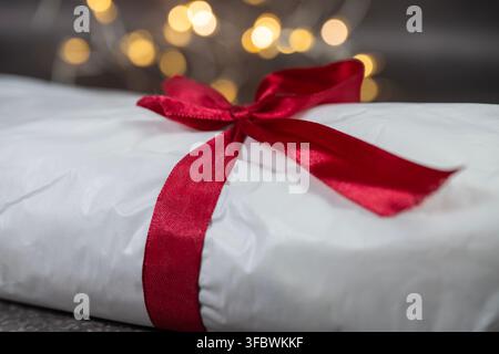 Un primo piano di un lungo pacco avvolto in carta bianca e legato con un fiocco di raso rosso festoso su uno sfondo scuro con luci sfocate a bokeh. Foto Stock