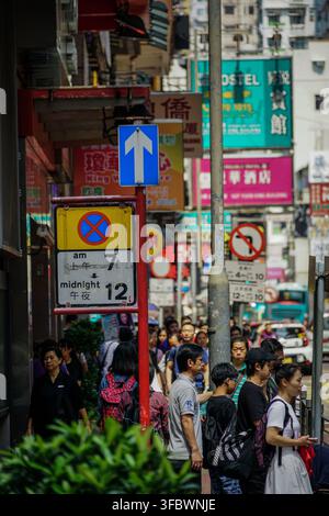 Una strada trafficata a Hong Kong presenta un mix di pedoni che camminano sul marciapiede, vari segnali stradali tra cui una freccia blu a senso unico e un cartello giallo che proibisce- Foto Stock