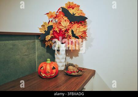 Decorazione autunnale per la casa su bancone di legno: vaso con foglie d'acero arancioni, decorazione di zucca jack-o'-lantern e piccola statuetta del bosco contro il muro bianco, accogliente s Foto Stock