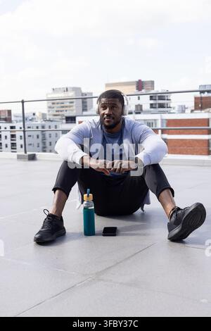Uomo africano di mezza età seduto sul tetto in abbigliamento sportivo, con cuffie e una bottiglia d'acqua nelle vicinanze Foto Stock