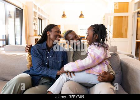 Famiglia afroamericana che sorride e si abbraccia su un divano grigio nel soggiorno con lampade a sospensione Foto Stock