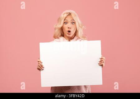 Giovane donna sorpresa che tiene un cartello vuoto con la bocca aperta, sfondo di studio, spazio per la copia Foto Stock