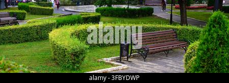 Panchina di legno vuota in un parco pubblico verde a Pyatigorsk, Russia. Paesaggio urbano con siepi curate, erba e vialetto lastricato. Infrastruttura da giardino cittadino Foto Stock