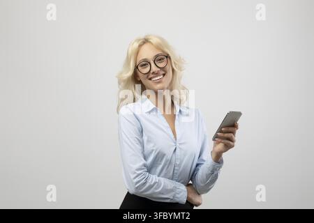 Giovane donna bionda sorridente che tiene uno smartphone in ufficio, concetto aziendale Foto Stock