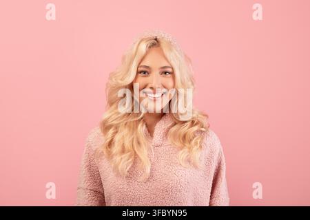 Giovane donna sorridente con capelli biondi in un maglione accogliente, sfondo rosa, concetto di bellezza Foto Stock