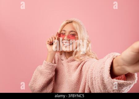 Giovane donna sorridente con occhiali che scatta un selfie a casa, sfondo rosa Foto Stock