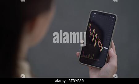 Trader donna monitora da vicino il prezzo di Bitcoin in tempo reale sul suo smartphone, analizzando grafici a candele rosse e verdi e il volume per le decisioni di trading. Foto Stock