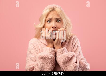 Giovane donna preoccupata con capelli biondi che tiene le mani sul viso, sfondo rosa Foto Stock