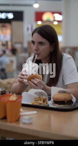Giovane donna in maglietta bianca sorseggia una bevanda attraverso una cannuccia mentre tiene un hamburger morso su un vassoio di fast food con patatine fritte in una food court affollata di un centro commerciale. Foto Stock