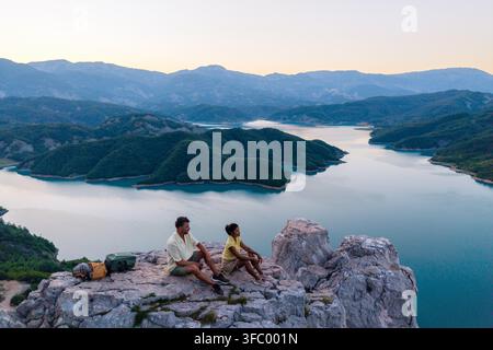 Due individui sono seduti su una sporgenza rocciosa, godendosi la vista mozzafiato sul tranquillo bacino artificiale di Bovilla in Albania. Il sole che tramonta getta tonalità calde sul paesaggio pittoresco. Foto Stock