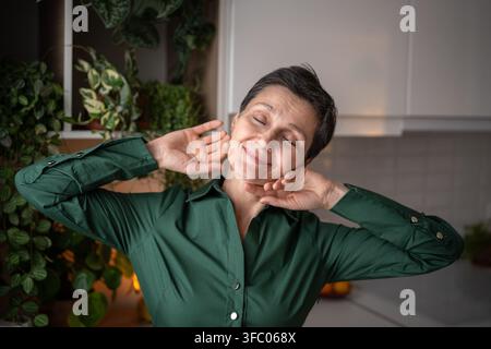 Una donna matura felice e soddisfatta che si gode la luce del sole mattutino nell'accogliente cucina casalinga durante i fine settimana Foto Stock