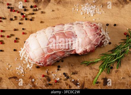 Vista dall'alto di arrosto crudo di spalla di maiale legato in una rete per carne su un tagliere di legno con spezie e rosmarino Foto Stock