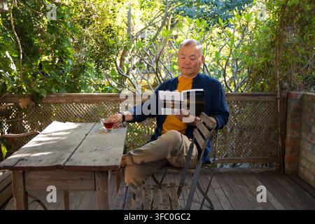 Uomo asiatico anziano seduto sul terrazzino che legge un libro tascabile mentre appoggia una tazza da tè in vetro sul tavolo Foto Stock