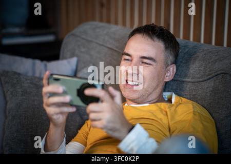 Giovane uomo felice che si rilassa su un divano a casa, ride guardando video divertenti o giocando a un gioco mobile sul suo smartphone. Intrattenimento, tempo libero an Foto Stock