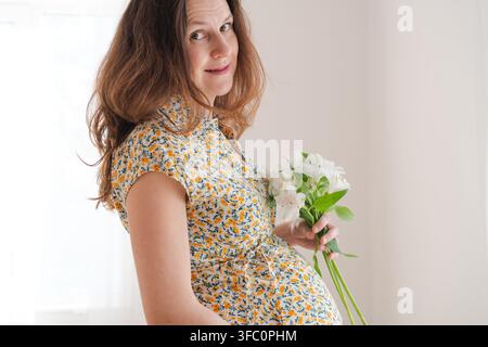 Una donna incinta in un vestito a fiori tiene un mazzo di fiori con un'espressione felice e allegra all'interno. La madre che gode dell'attesa mostra un calore materno splendente e gioioso in un interno luminoso. Foto Stock