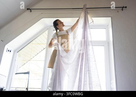 La donna in grembiule appende tende in tulle trasparente su grandi finestre sottotetto nella casa all'interno dell'interno bianco. Pulizia di primavera, rimessa in ordine Foto Stock