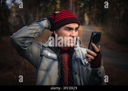 Uomo che si tiene la testa in stato di shock mentre guarda il suo smartphone in un ambiente boschivo, sentendosi perso. Segnale perso, nessuna connessione e bloccato nel bosco Foto Stock