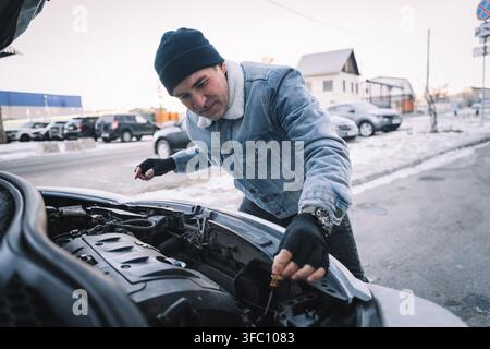 Uomo che controlla il livello dell'olio motore in un'auto durante l'inverno, tenendo una bacchetta indicatrice, eseguendo la manutenzione e l'ispezione del veicolo Foto Stock