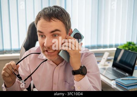 Uomo d'affari arrabbiato che parla al cellulare in un ufficio, mostrando frustrazione e confusione durante una conversazione o negoziazione commerciale tesa. Foto Stock