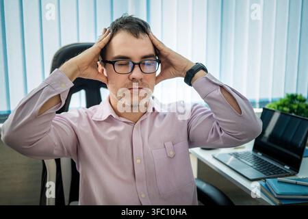 Dipendente d'ufficio stressato che tiene entrambe le mani sulla testa per la frustrazione mentre è seduto alla sua scrivania. Forte mal di testa, esaurimento mentale e pressione sul lavoro Foto Stock