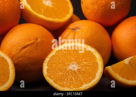 Un mucchio di arance intere fresche, arance tagliate a metà e un quarto sono disposti su uno sfondo scuro. Foto Stock