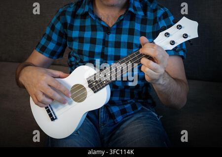 Primo piano di un uomo in una camicia a quadri blu che suona un ukulele bianco seduto su un divano grigio. Foto Stock