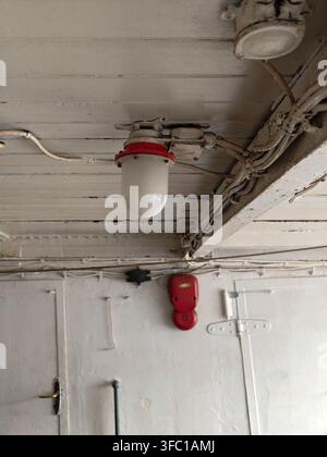 Un'inquadratura angolata verso l'alto che cattura una lampada in stile industriale con base rossa e globo bianco, montata su un soffitto in legno bianco stagionato. Elettricità esposta Foto Stock