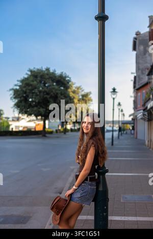 Giovane donna che posa accanto a un lampione con cappello da cowboy. Giovane donna dai lunghi capelli castani che posa accanto a un lampione in pantaloncini di jeans e top marrone mentre Foto Stock