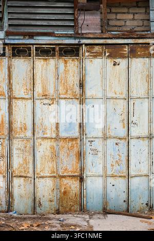 Porta pieghevole in metallo consumata con vernice blu scrostata, ruggine pesante, vecchie persiane in legno e muratura in mattoni esposta su un edificio antico a Bukit Mertajam, Penang Foto Stock