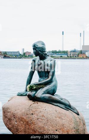 Copenaghena, Danimarca - 13 maggio 2026: Una vista verticale della statua della Piccola Sirena a Copenaghen mostra la sua superficie di bronzo bagnata, lo sguardo abbassato e una bouque Foto Stock