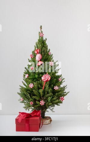 Piccolo albero di Natale decorato con ornamenti alla moda di caramelle rosse accanto a una scatola regalo rossa su sfondo bianco. Decorazione festiva per le vacanze di Natale. Formato verticale. Foto Stock