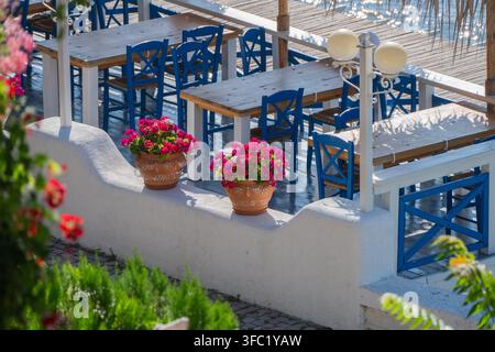 Un'incantevole terrazza di caffè all'aperto in stile mediterraneo presenta muri imbiancati a calce, tavoli in legno rustico, sedie blu classiche e vasi di terracotta pieni Foto Stock