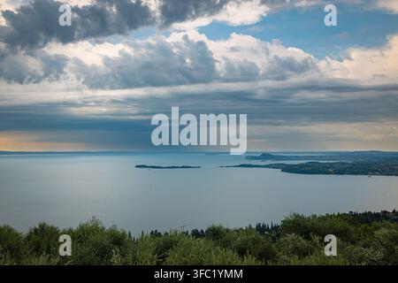 Panorama sereno del lido con isole sotto un cielo nuvoloso drammatico sopra le acque calme del Lago di Garda, Italia Foto Stock