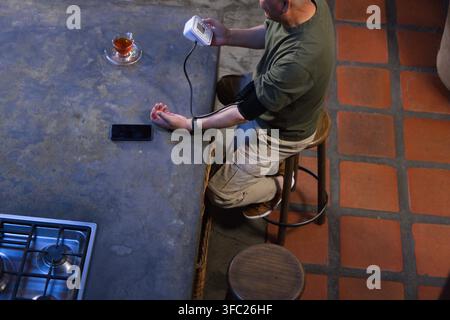Uomo anziano seduto su uno sgabello di legno al bancone della cucina che misura la pressione sanguigna con un monitor digitale Foto Stock