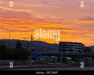Un drammatico alba infuocata dipinge il cielo di arancione sopra un moderno edificio residenziale, una strada e le montagne, catturando una tranquilla mattina urbana. Foto Stock