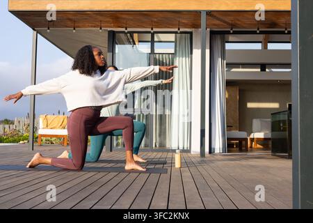 Diverse partner femminili che praticano yoga su un terrazzo di legno con leggings marrone, usando materassini e una bottiglia d'acqua Foto Stock