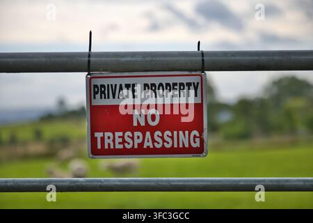 Insegna rossa e bianca con la scritta "Proprietà Privata Divieto di Accesso" attaccata a un cancello di metallo davanti a un campo verde rurale con pecore. Foto Stock