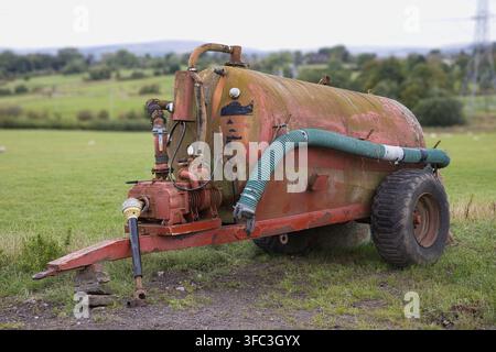Rimorchio cisterna per liquami rosso vintage parcheggiato in un campo agricolo verde, con vernice scolorita, ruggine e un pesante tubo di aspirazione. Foto Stock