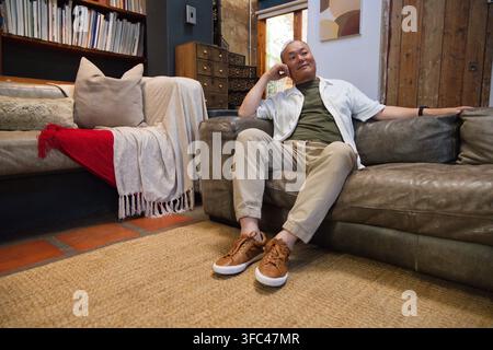 Uomo asiatico anziano seduto su un divano di pelle marrone, appoggiato all'indietro, guarda a sinistra nel soggiorno Foto Stock
